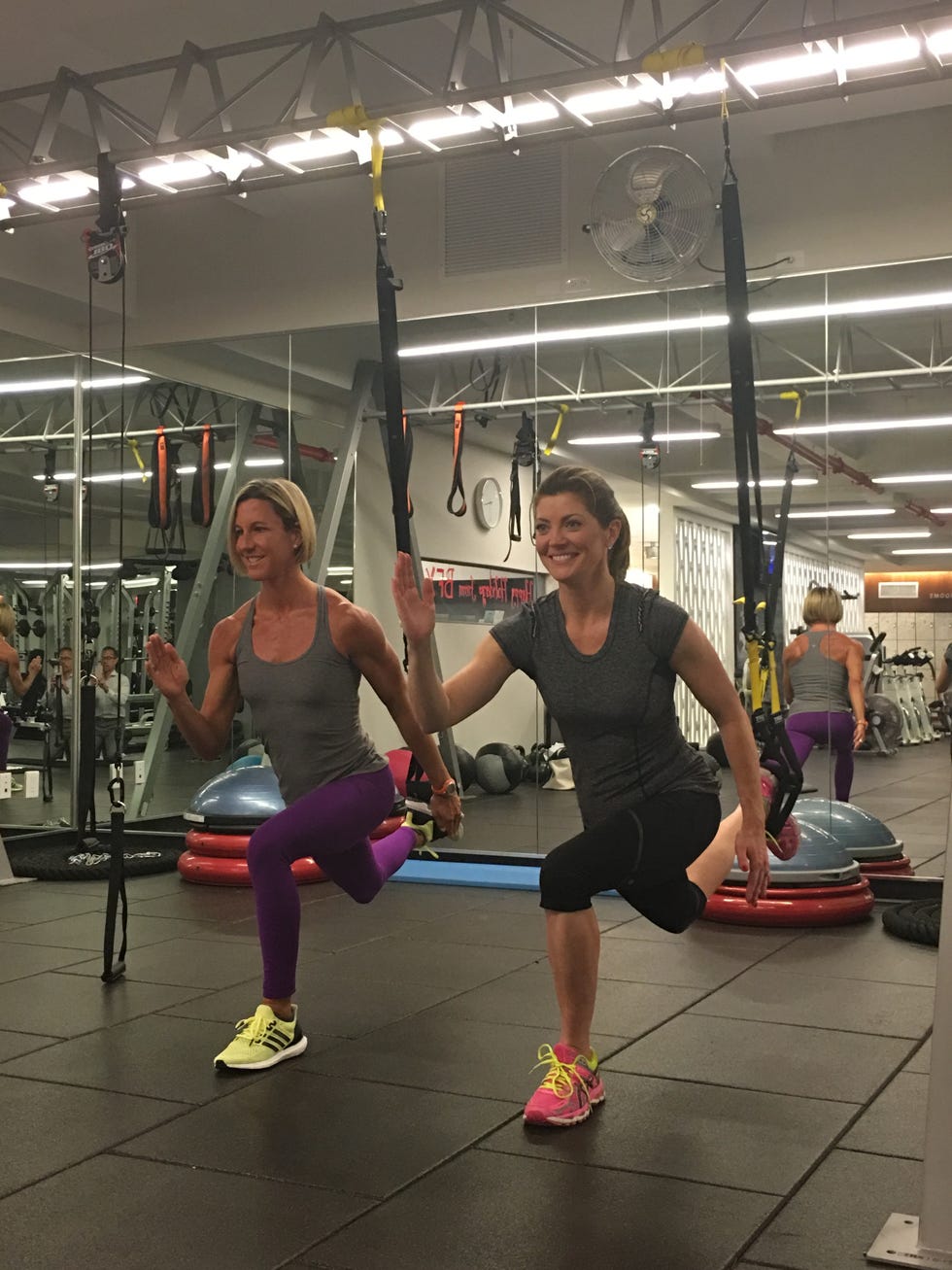 Two women performing fitness exercises together in a gym, using resistance bands for support and balance.