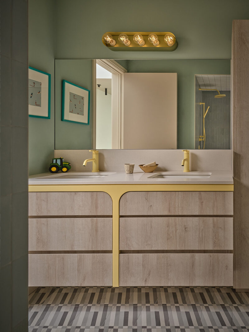 Bathroom in a Gillian Gillies Net Zero Ready Home with dual sinks, light wood vanity, yellow accents, large mirror, wall-mounted lights, green walls, framed art, and a toy tractor on the counter.