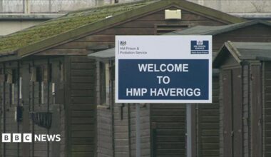 A blue-and-white sign saying "Welcome to HMP Haverigg". It is in front of hut-style buildings painted brown outside the main gate of the prison.