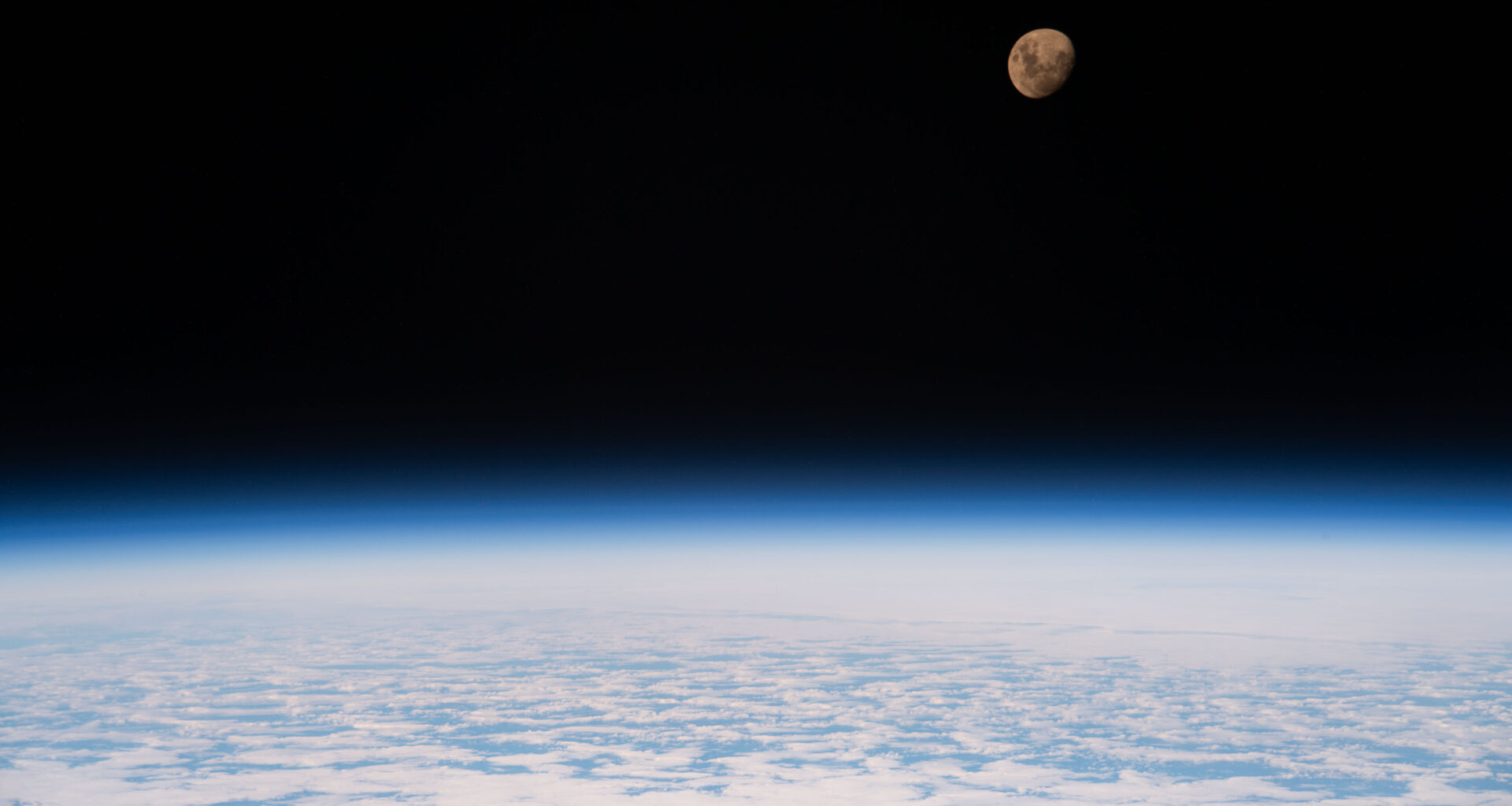 A view of the Moon from the International Space Station as it orbited 264 miles above a partly cloudy Indian Ocean southeast of Madagascar. The Moon has a golden glow to it, appearing more bronze than its usual gray. It stands out against the darkness of space, while below, Earth's atmosphere glows blue. Below that, we can see white clouds covering up most of the water below.
