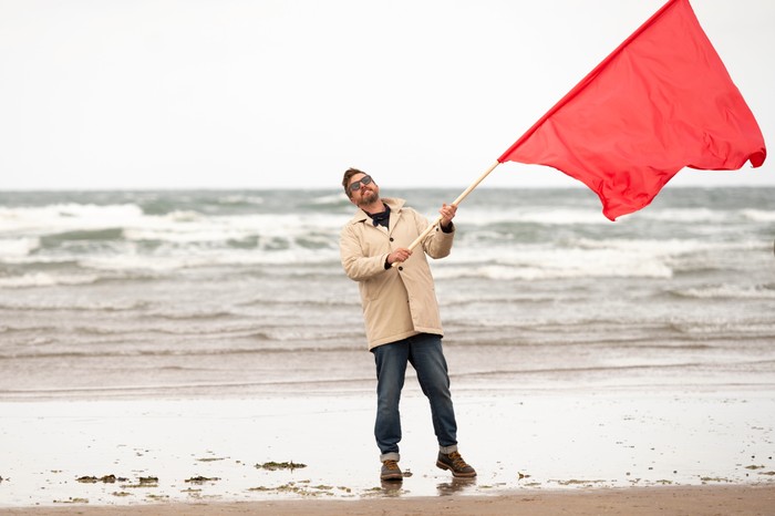 571934,TITLE:Race Against the Tide Iain Stirling stood on a beach waving a large red flag.