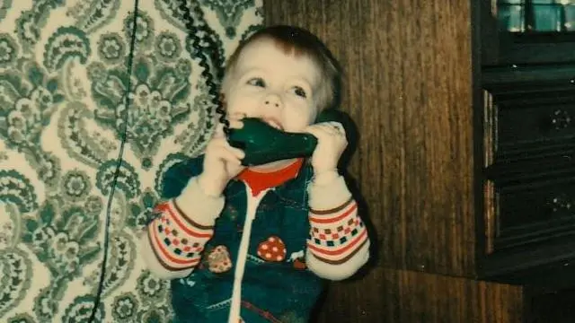 A vintage photograph show Nina at three years old, playing with an old fashioned rotary style black phone. She dey look up and smile. Di room get a distinctive 1970s patterned wallpaper and dark furniture.