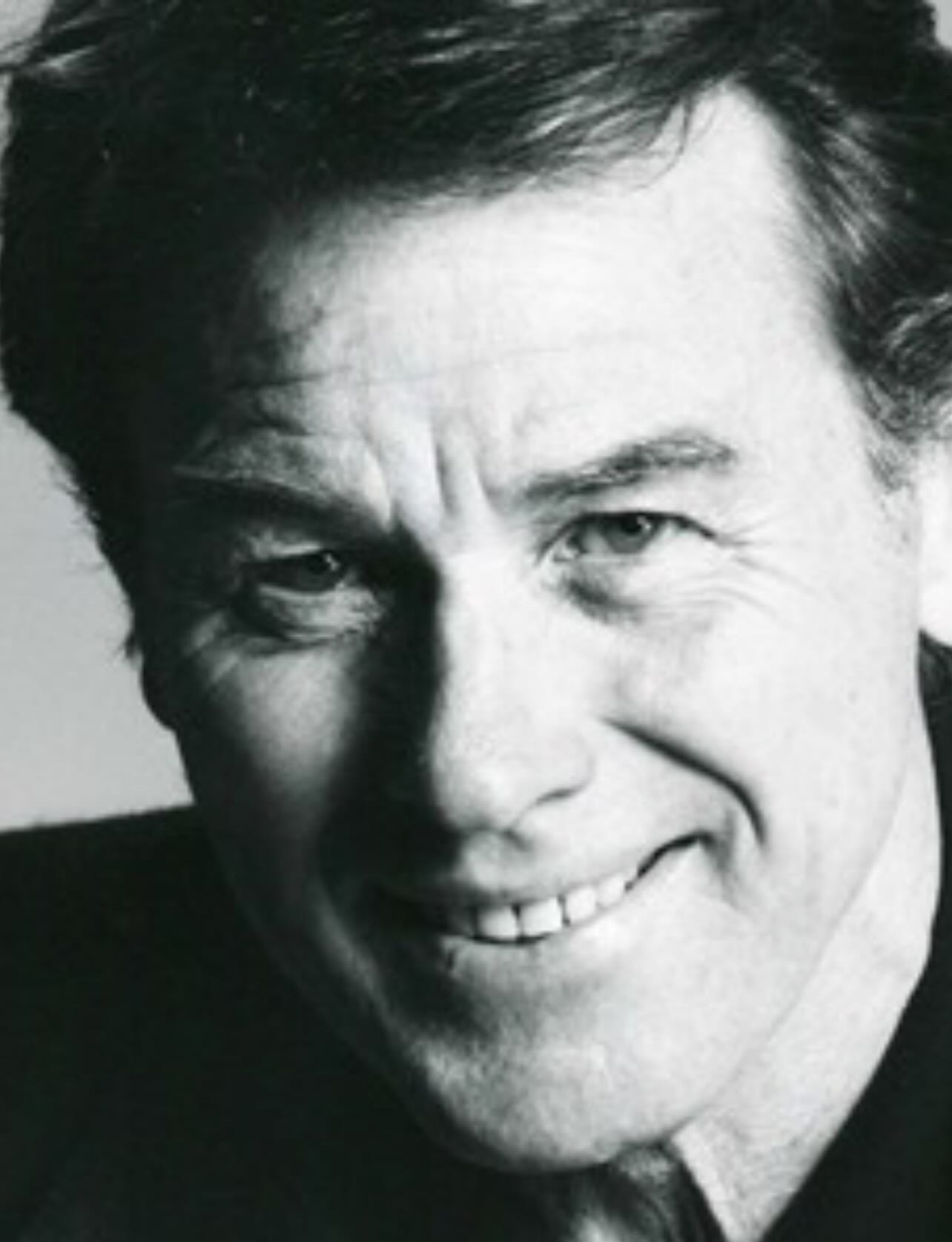 A close up black and white photo of a man smiling while looking at the camera.