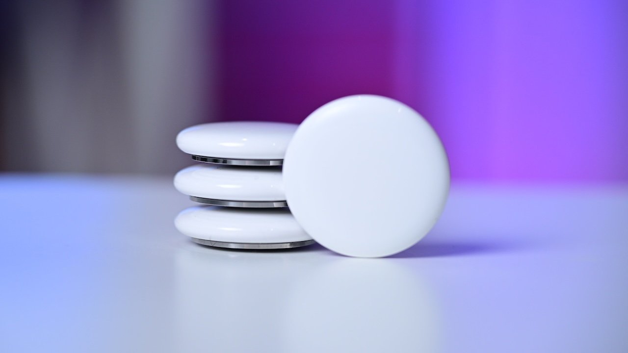 Stack of Apple AirTag trackers on a smooth surface, one standing upright in front, with a blurred purple and blue background