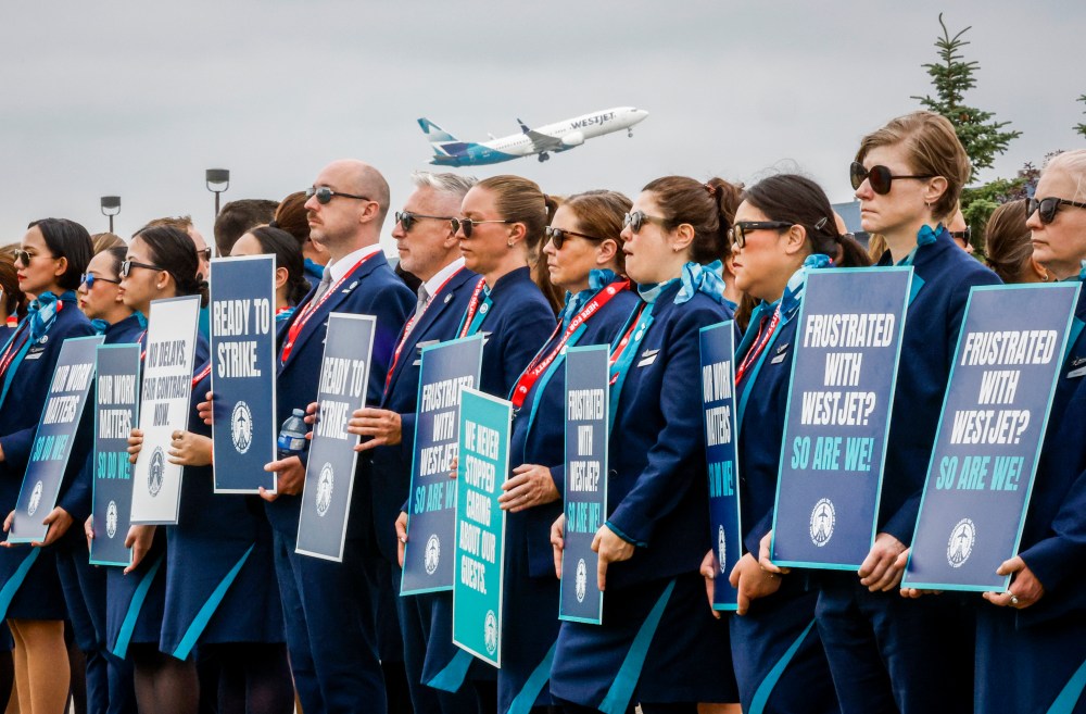 A WestJet plane takes off as WestJet flight attendants hold a