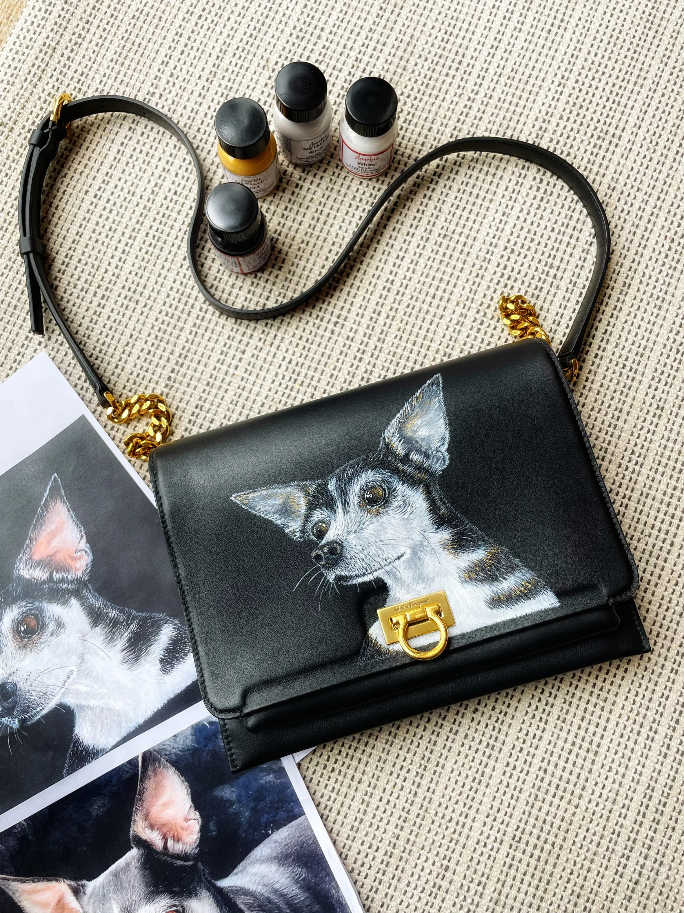 A black leather handbag with a painted portrait of a black, white, and brown dog, next to tubes of gold and silver paint.