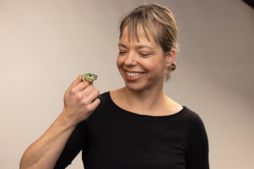 A woman in a black shirt with blonde hair holds a small green frog on her hand.