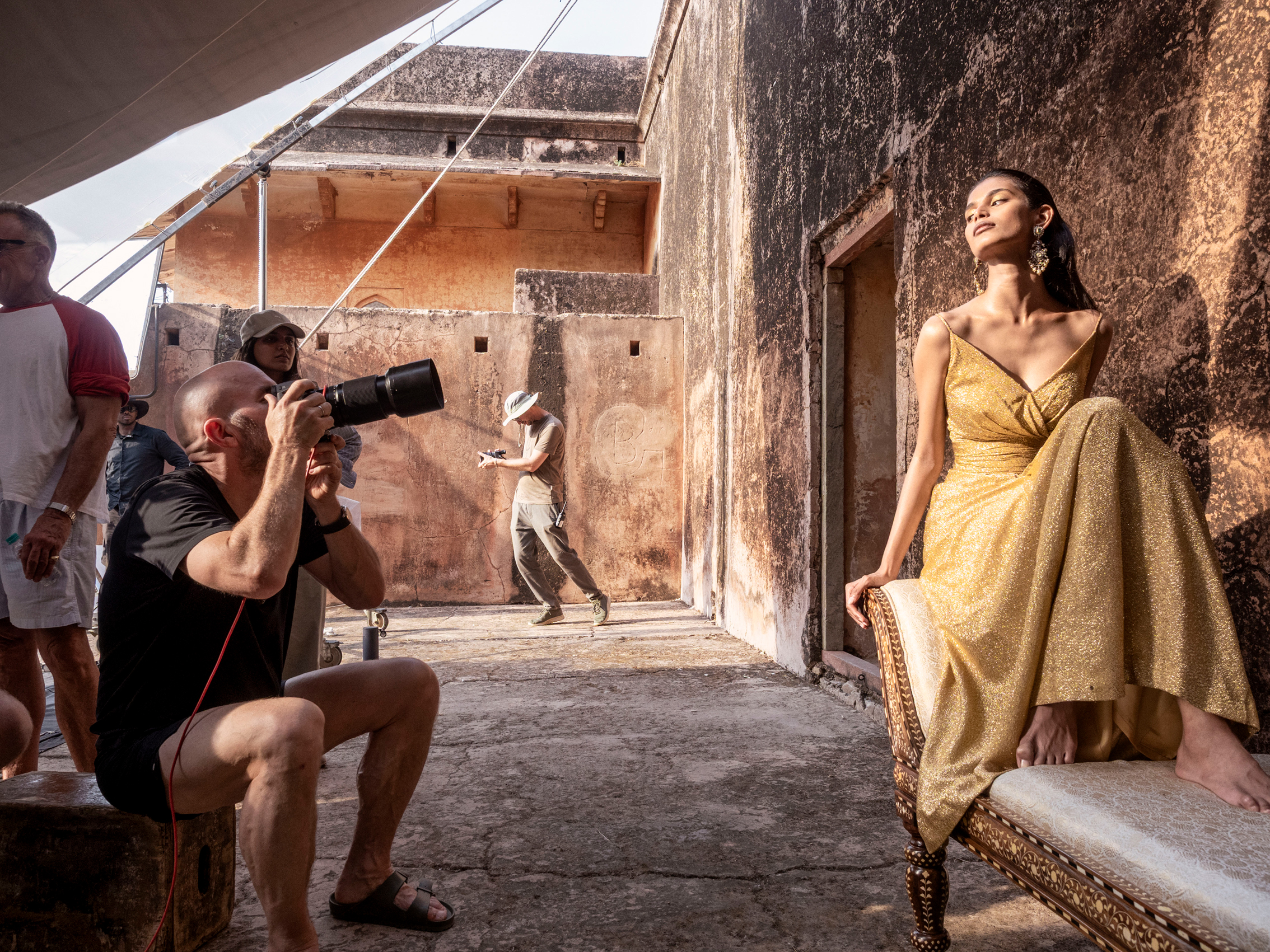 A cream colored backyard, left a woman with long dark hair posing in a golden dress on a baroque style sofa, left the photographer pointing a big lens towards her
