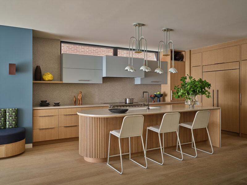 Modern kitchen designed by Gillian Gillies in a Net Zero Ready Home, featuring light wood cabinets, a curved island with three white barstools, blue accent wall, and pendant lights. A plant and decorative items are arranged on the counters and shelves for a stylish, sustainable ambiance.