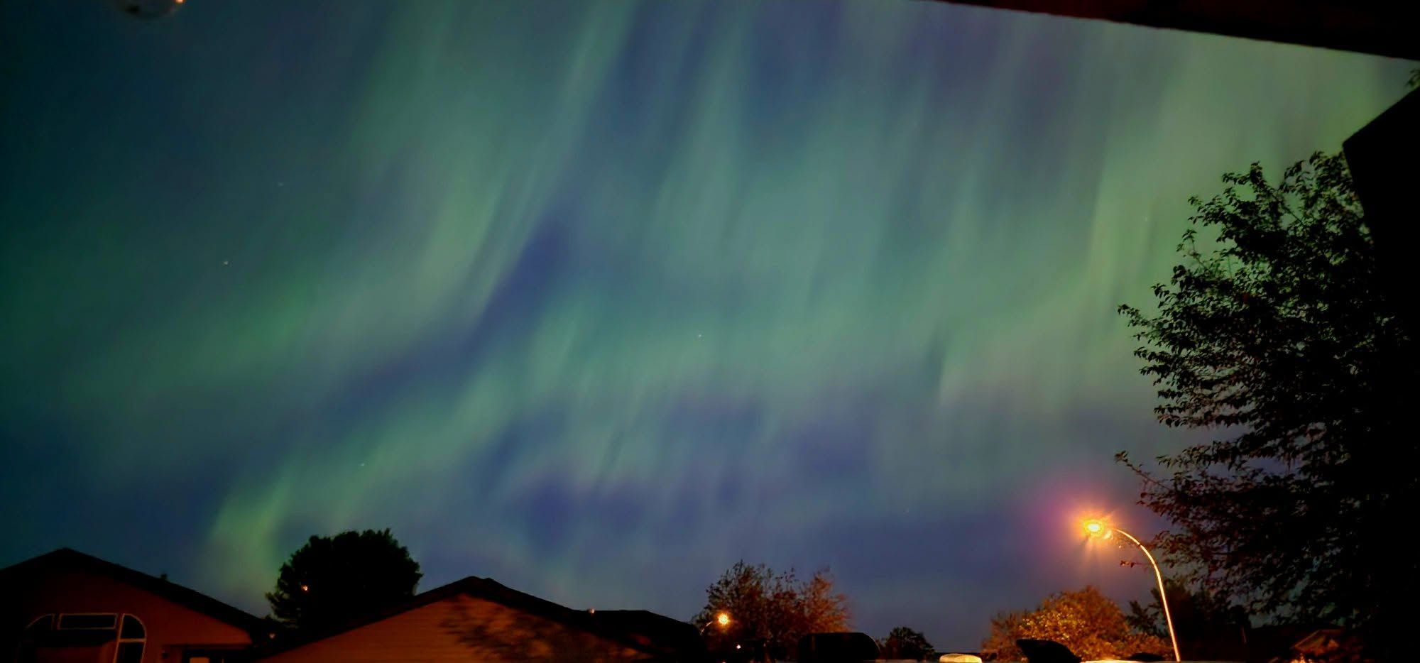 Flowing green ribbons of the aurora borealis sweep across the night sky above a residential neighbourhood, creating a vivid display of northern lights over the rooftops below. Photo Lisa Marie