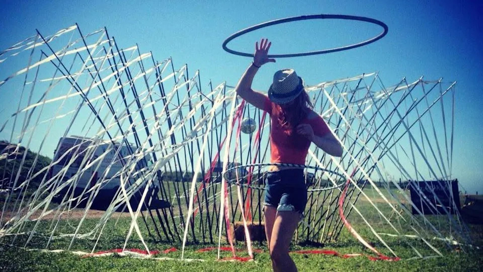 Lowri dancing with a hula hoop around her waist. She is wearing a red short-sleeved top and denim shorts and in a field with an art installation covered in streamers behind her.