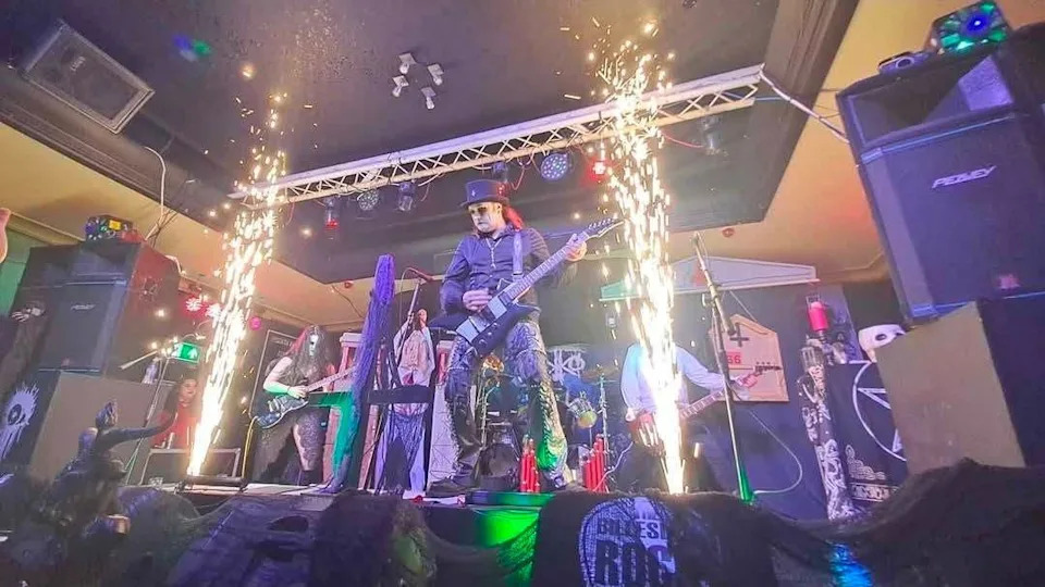 A man is wearing an all black outfit and a top hat. He is holding a guitar with band members standing behind him as sparklers spray from the stage.