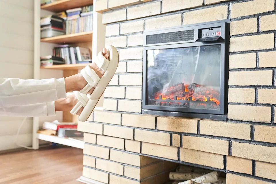 Person wearing sandals relaxes by a brick fireplace, warming their feet. Bookshelves visible in the background. Cozy, indoor setting