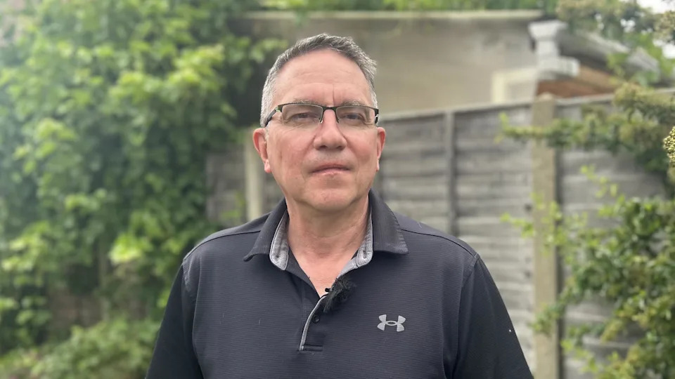 A man with short grey hair is wearing glasses and a black polo shirt while standing in front of a garden fence.