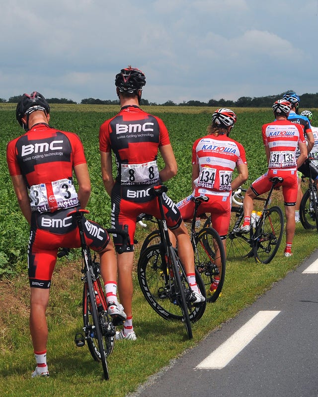 Cyclists with BMC jerseys paused on the roadside during a race, surrounded by support vehicles and green fields. Cyclists with BMC jerseys paused on the roadside during a race, surrounded by support vehicles and green fields.