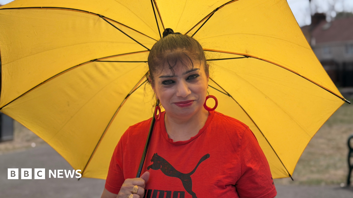 Amelia Lord is a white woman in her late 20s. She has shoulder length brown hair partly pulled back in a ponytail with frontpieces either side of her face. She has defined eyebrows and is wearing makeup, has a central nose ring and earrings, and is smiling at the camera. She wears a sleeveless black top. She is holding a pair of books and stands in front of a bookshelf with collections of books on it, including titles by Rebecca Yarros and the Harry Potter series by JK Rowling.