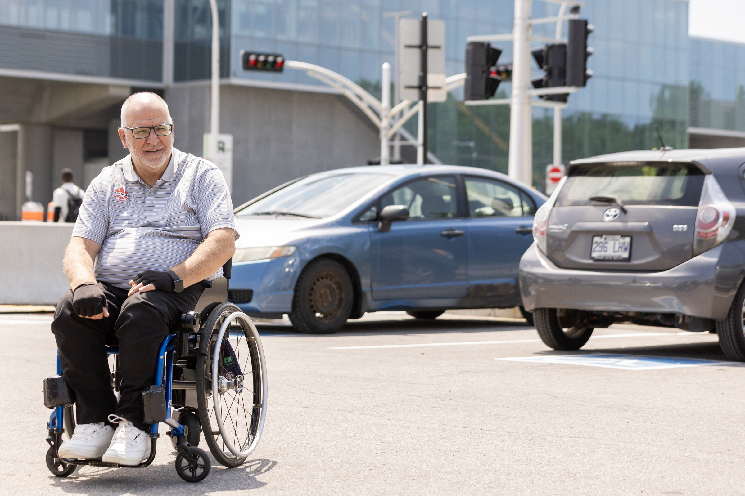 A man in a wheelchair with a pair of accessible parking spaces behind him. A REM station is visible across the street.