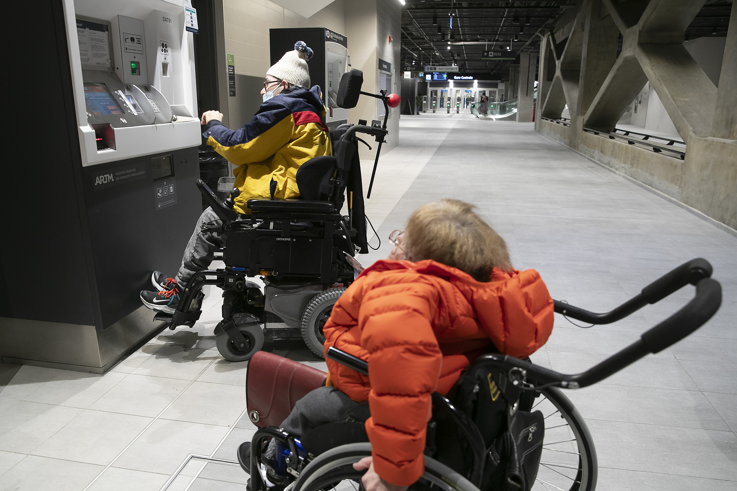 Two people in wheelchairs next to an automated machine whose screens angle upwards above their heads.