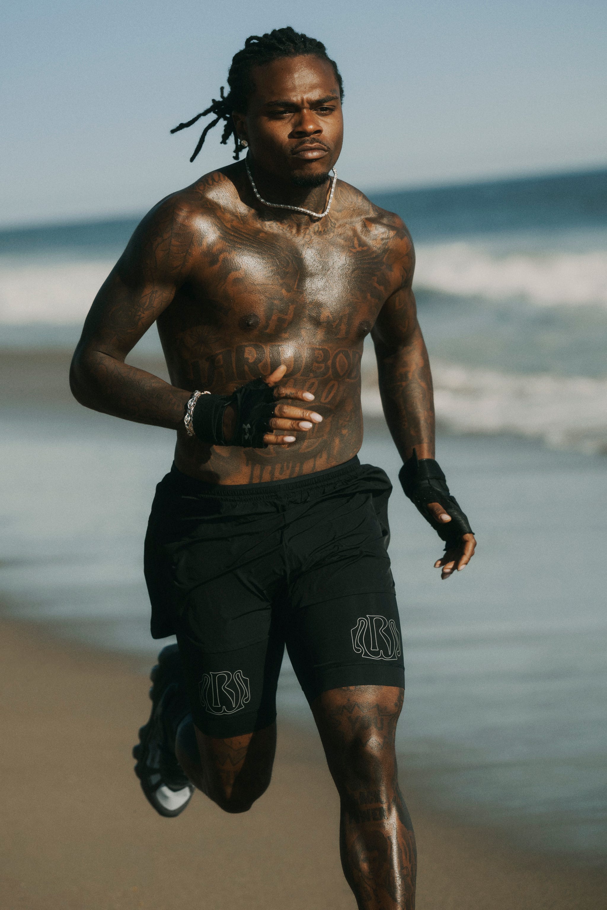 Athletic individual jogging along the beach with visible tattoos and wearing black shorts and gloves.