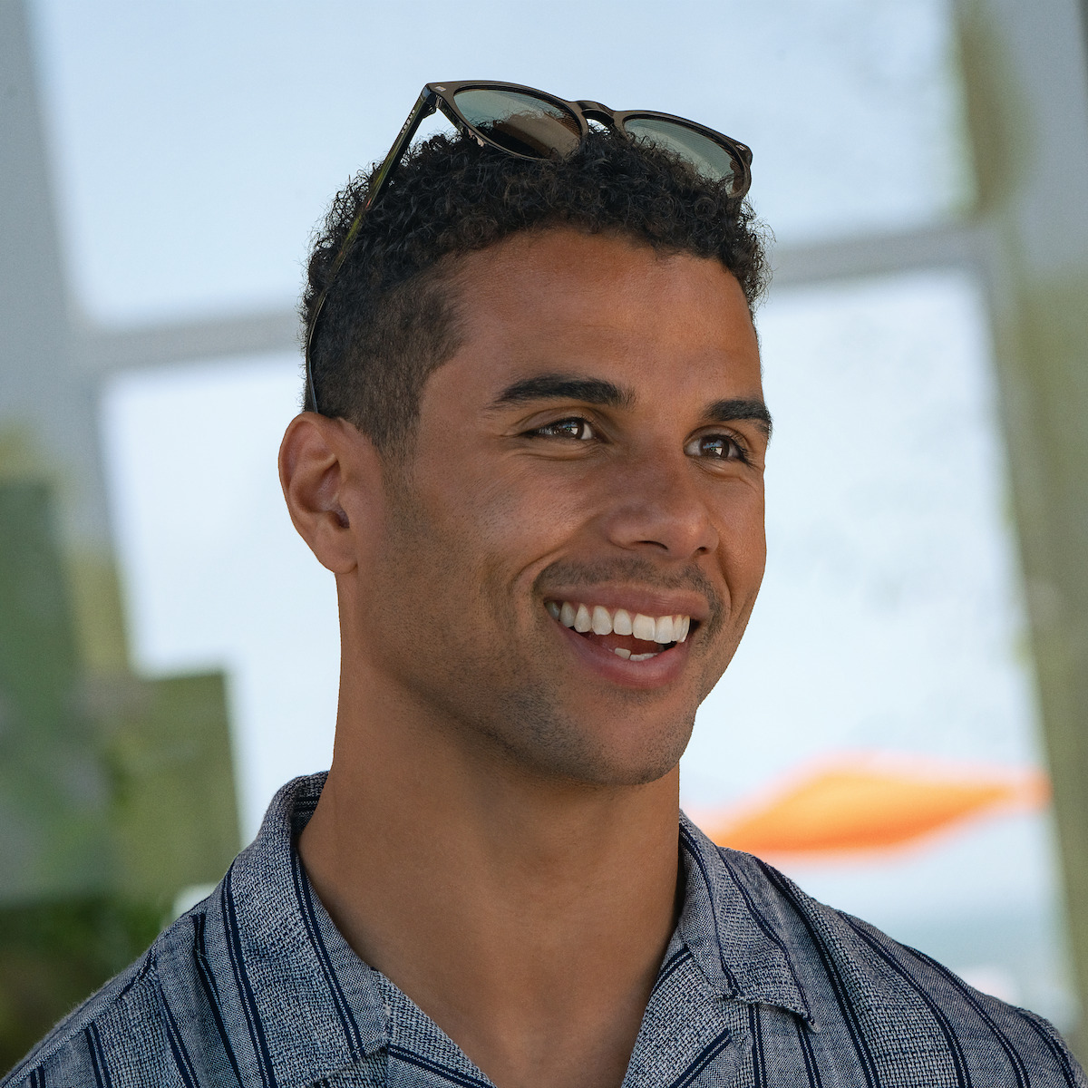 Mason Gooding as Mason with short curly hair and sunglasses on his head, wearing a striped shirt. He is indoors with sunlight and blurred outdoor scenery, possibly a beach or waterfront, visible through windows behind him. Relaxed, casual setting.
