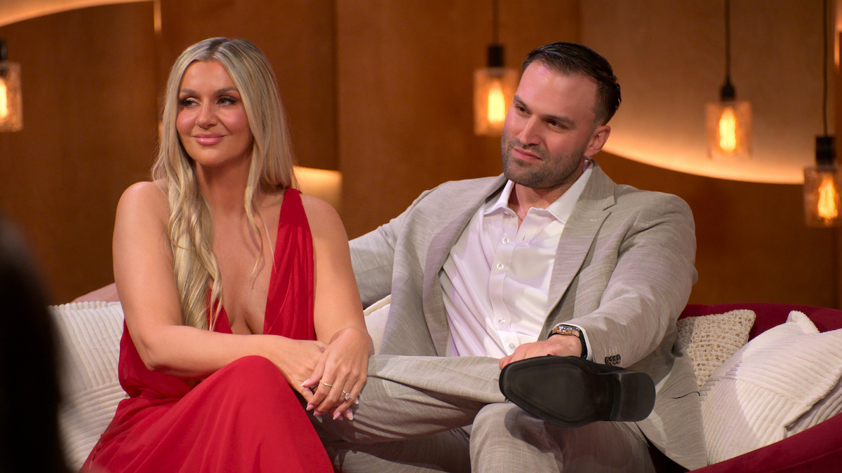 A woman in a red dress and a man in a light suit sit together on a sofa in a warmly lit, modern room with pendant lights and tan walls, appearing relaxed and engaged in conversation.