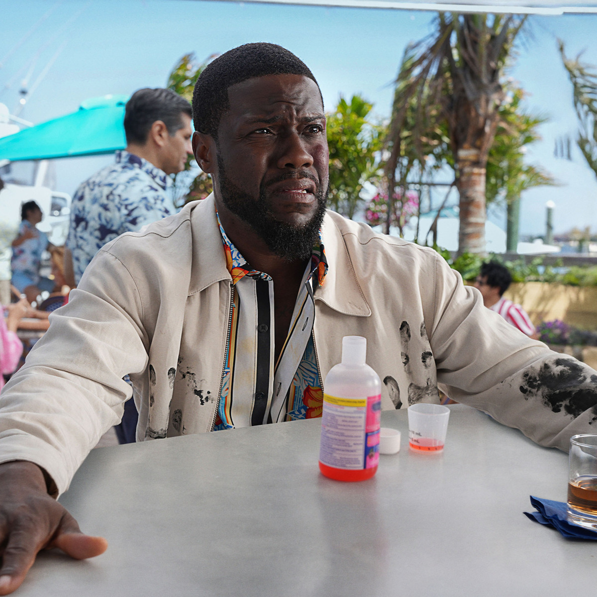 Kevin Hart as Joe with a concerned expression sits at an outdoor café table with a bottle and cup of medicine, surrounded by tropical plants, palm trees, and other people in casual attire.