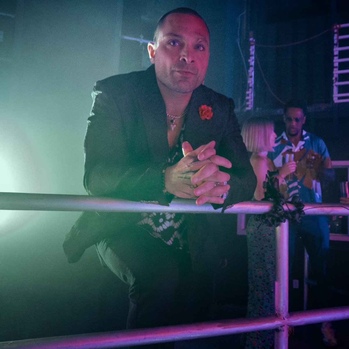 Michael Mando as Jaze in a black suit with jewelry leans on a railing in a dimly lit nightclub, with a floral lapel pin, while two people converse in the background among neon lights and industrial decor.