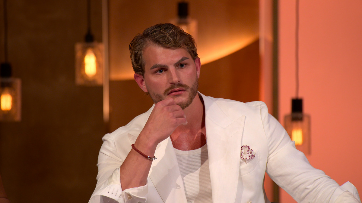 Man in a white suit sitting indoors, thoughtfully resting his chin on his hand, with warm hanging lights and brown-orange background.