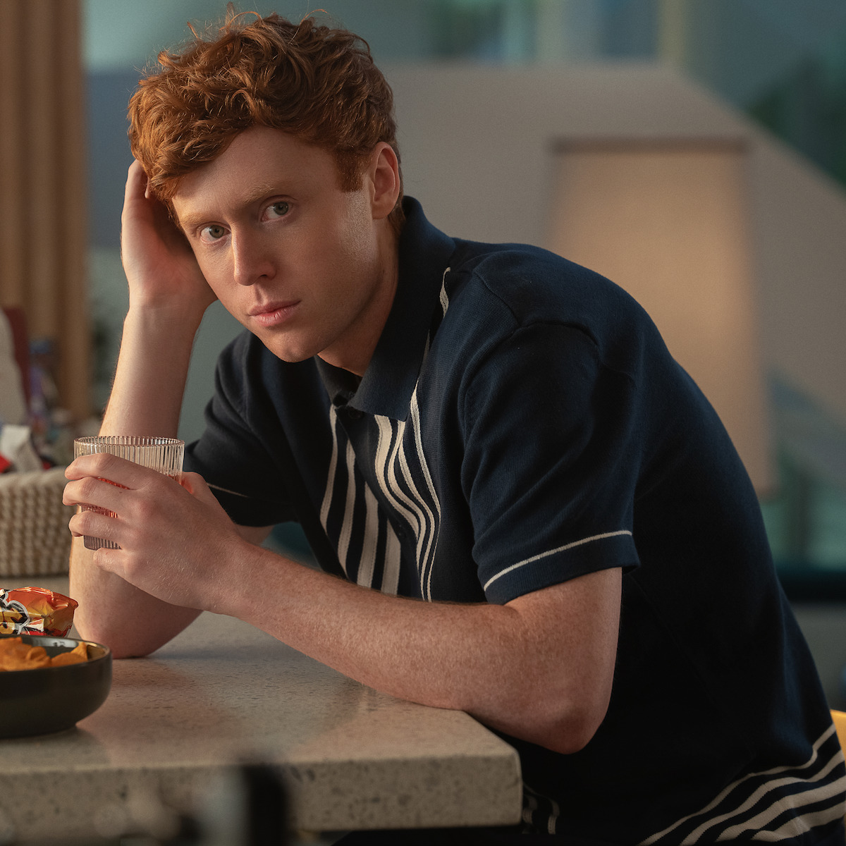Ben Marshall as Hunter with red hair sitting at a kitchen counter, holding a glass, looking at the camera with a relaxed expression; soft indoor lighting, casual setting with food and blurred background elements.