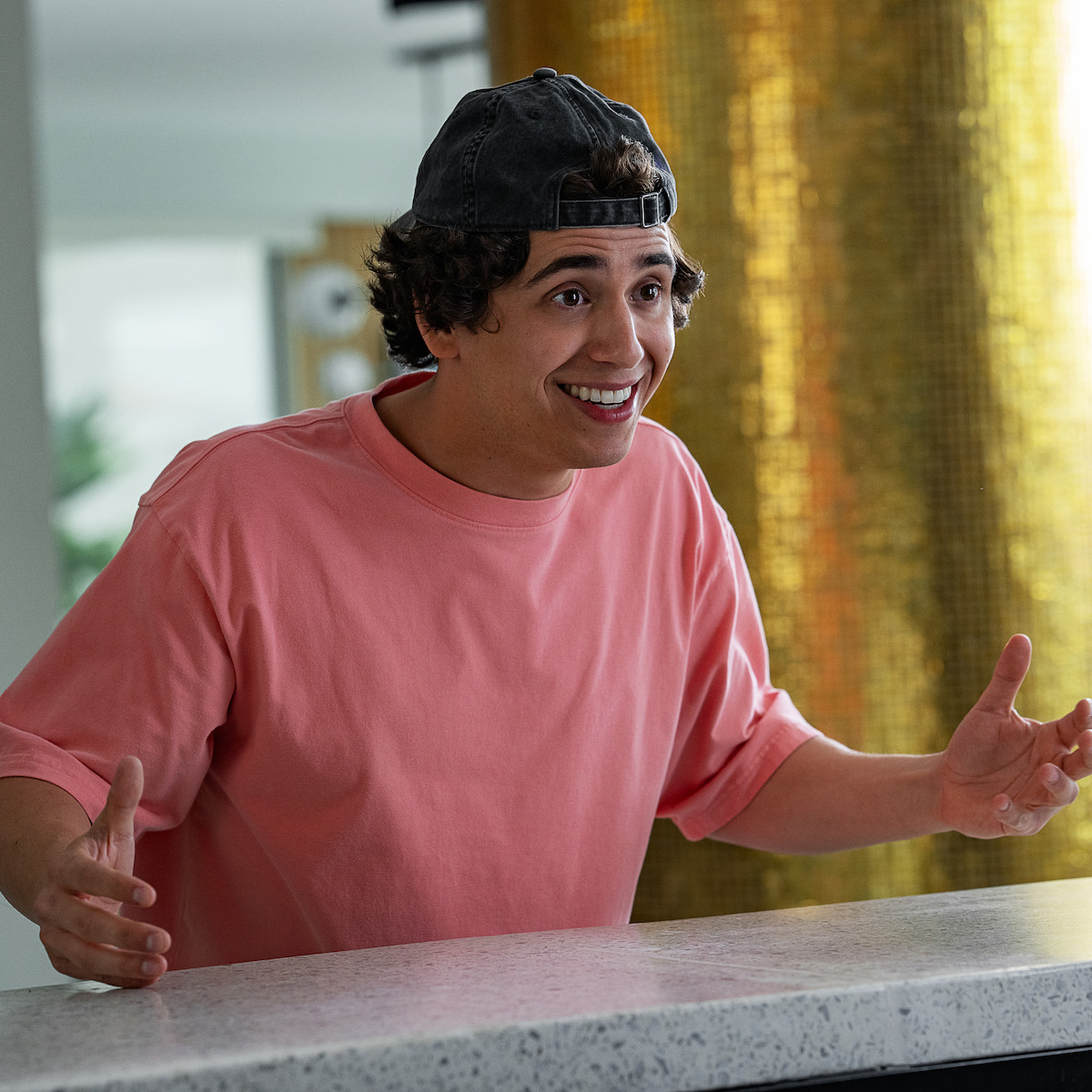 Marcello Hernandez as Nick in a pink shirt and backwards cap gestures energetically at a countertop indoors, with a gold-toned column and modern decor in the background.