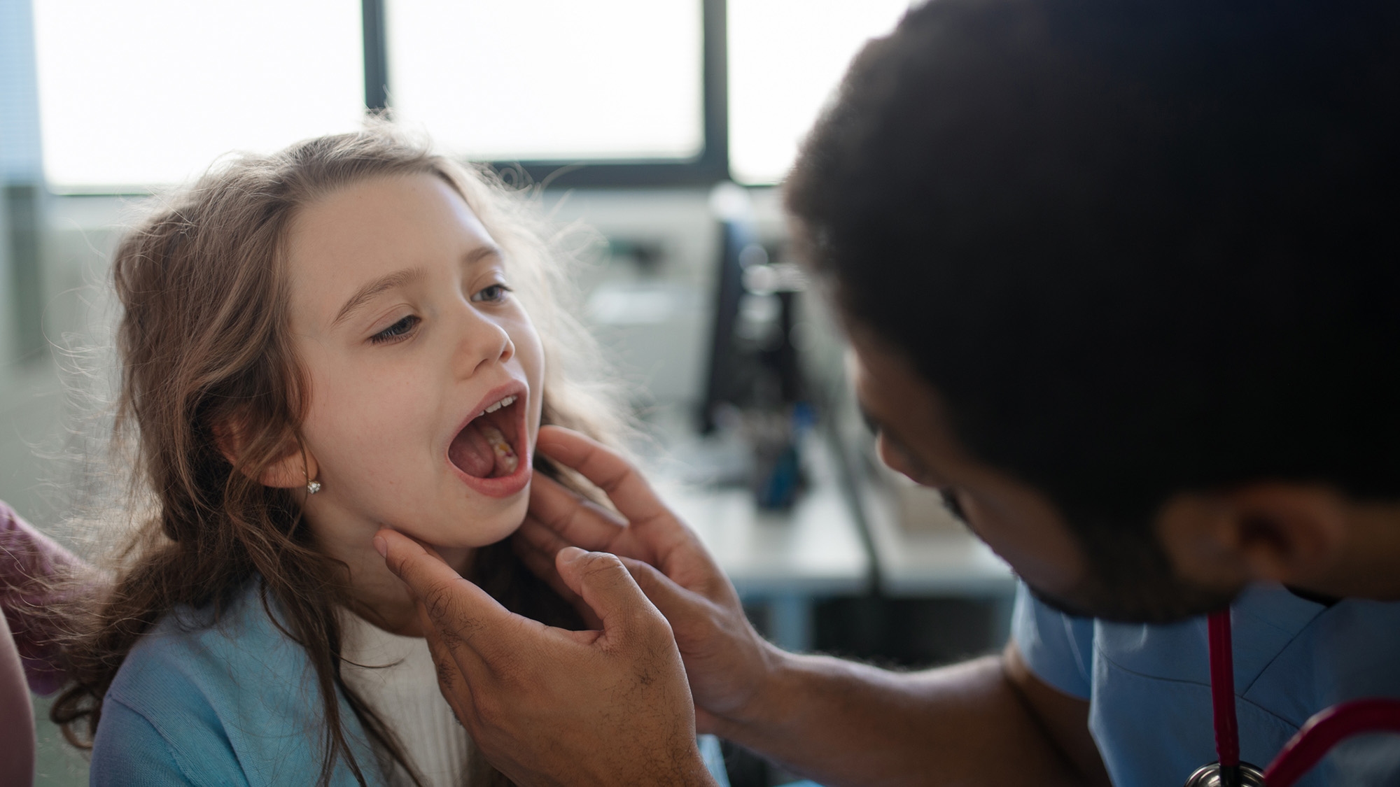 A young girl with long brown hair opens her mouth as a male nurse feels her lymph nodes.