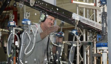 A man wearing ear protection works closely with multiple hydraulic and instrumentation units connected to a large beam mounted on a test structure. Numerous cables, hoses, and measurement devices extend from the setup.