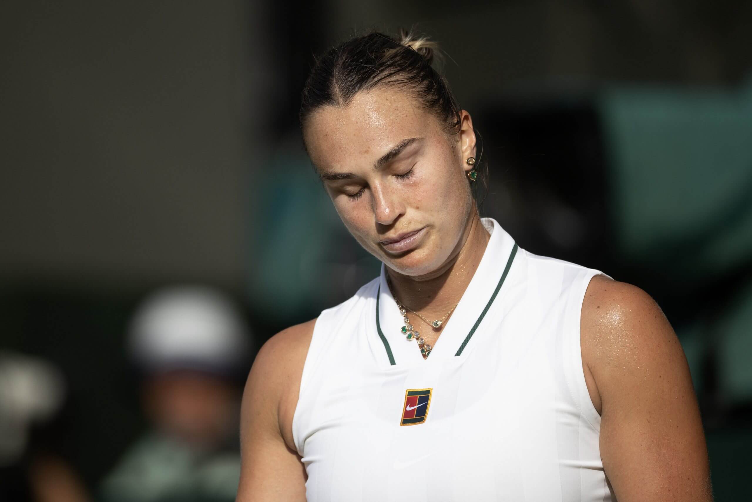 Aryna Sabalenka looks down despondently with her eyes closed, wearing a white tennis outfit.