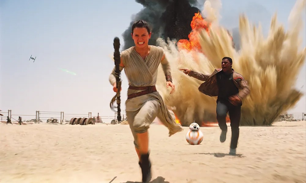 In a scene from The Force Awakens, Rey and Finn sprint across a desert landscape while escaping a massive explosion with BB-8 following closely behind them