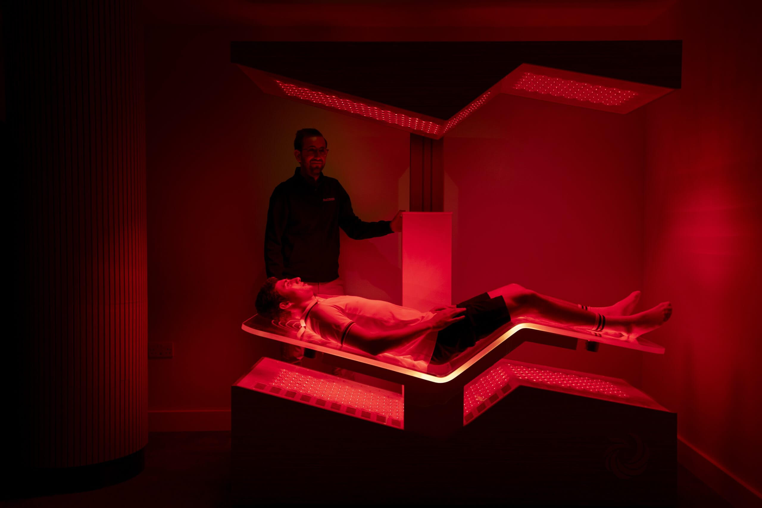 A red-light chamber with a bed in it, on which a man is lying while another man adjusts the settings.