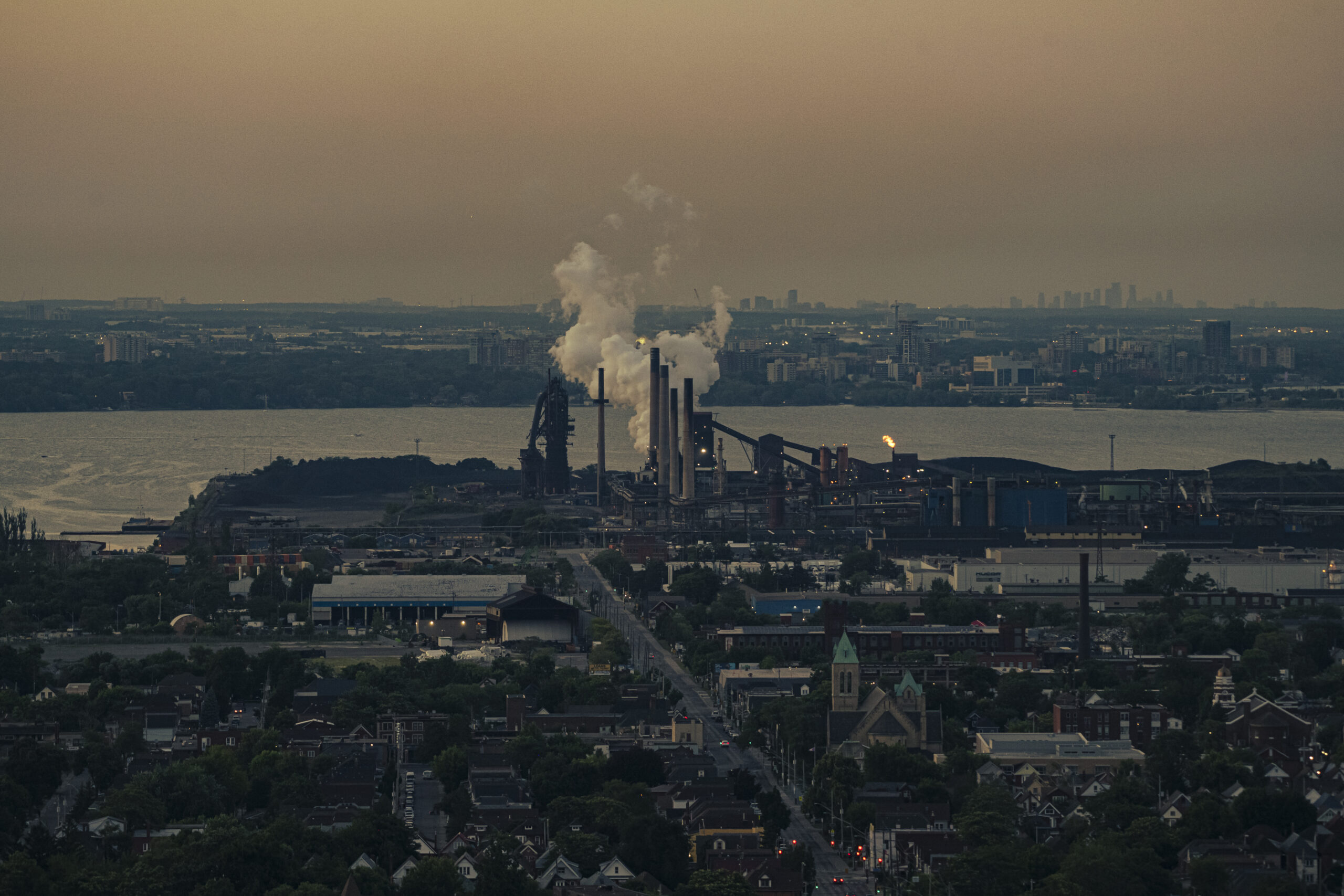 Smoke releases from stacks on a factory on the Hamilton Harbour, with Toronto's skyline in the distance