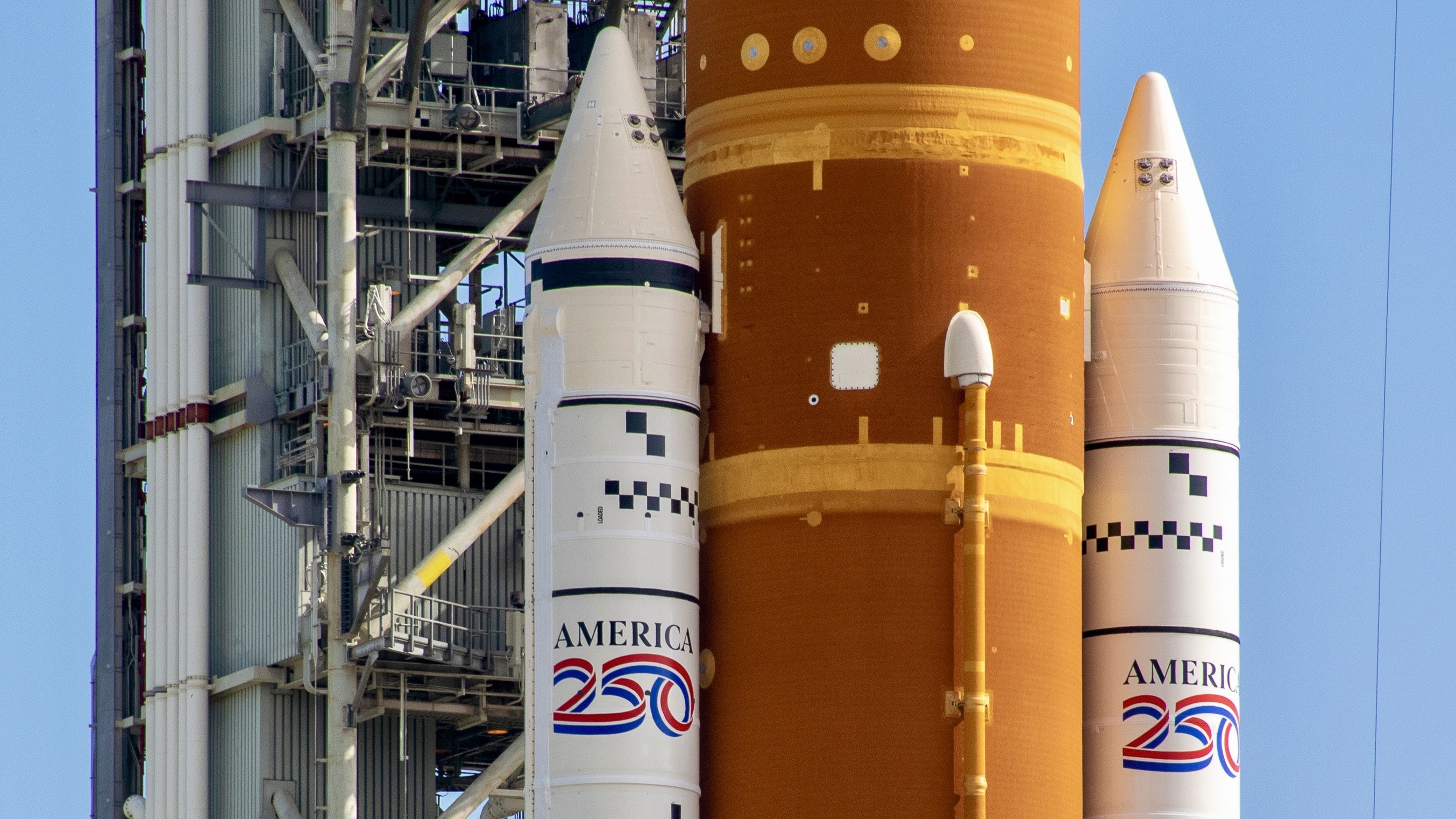 The mid section of an orange rocket with white side boosters stands against a metal tower. America 250 is decaled on both boosters.