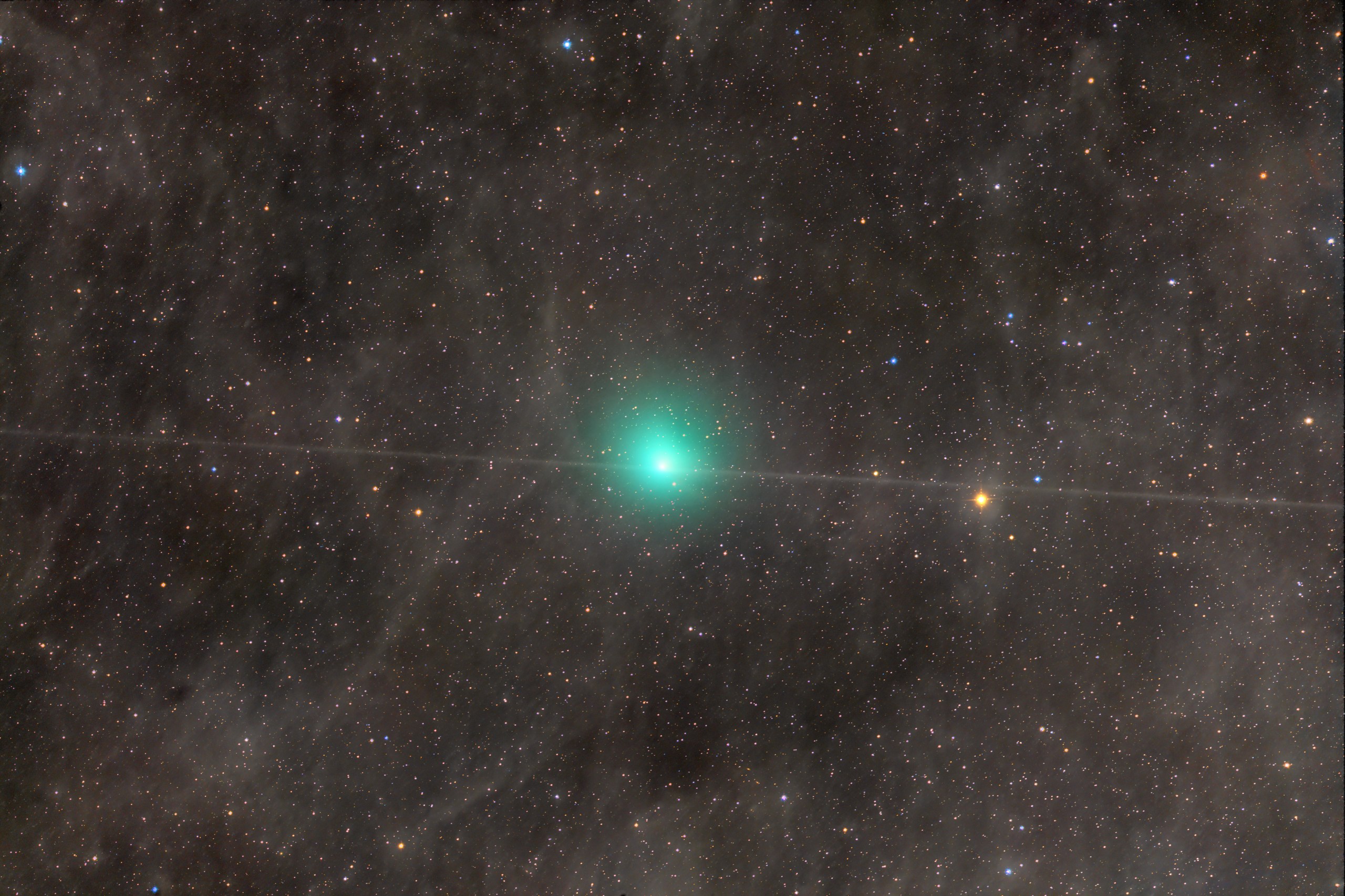 A telescope image of Comet 10P/Tempel 2, showing a bright blue-green coma glowing near the center of a richly star-filled field. A diagonal satellite or aircraft trail crosses through the frame. The surrounding star field is dense with colorful blue, orange, and white stars against a dark background.