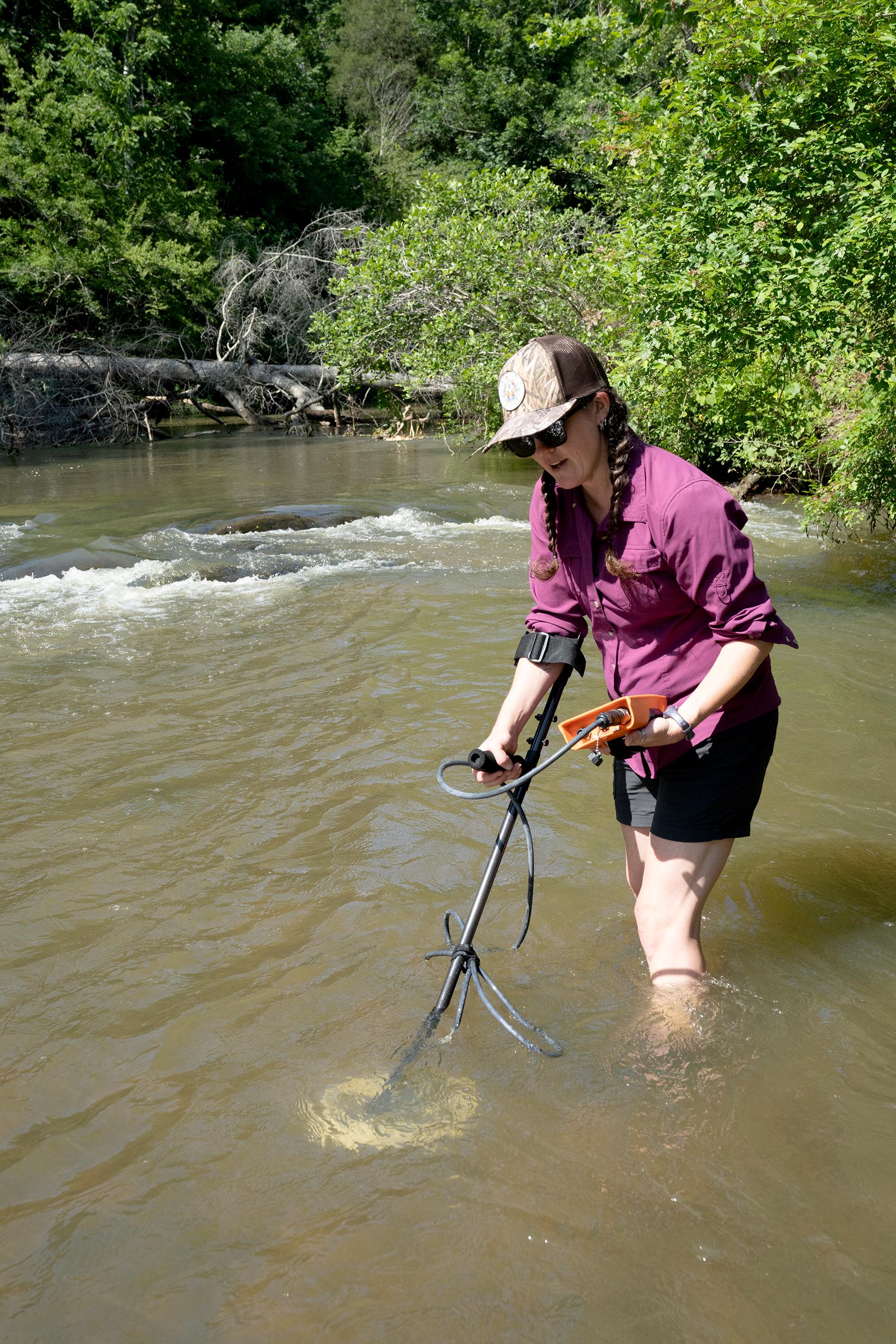 A woman in sunglasses, hat and shorts uses an electronic reader and detector to search through a stream.