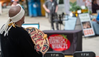A person viewed from behind plays an Indigenous hand drum in the foreground. A farmers market stall is out of focus in the background.