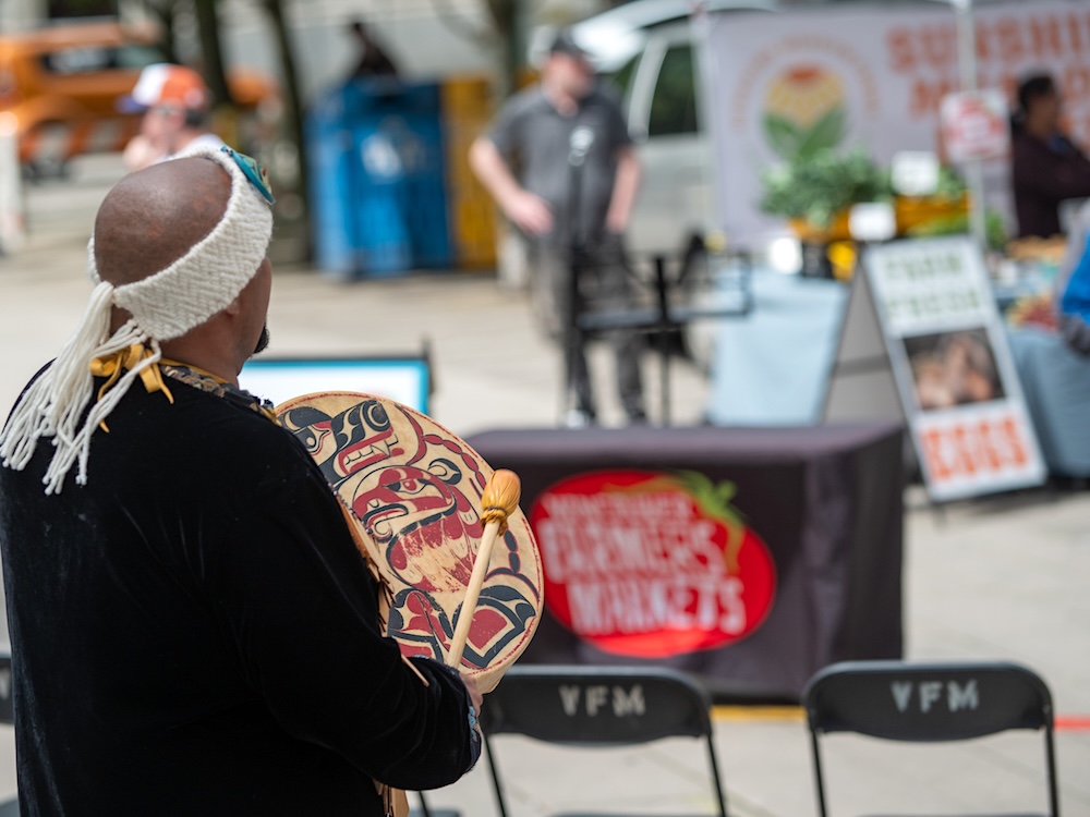 A person viewed from behind plays an Indigenous hand drum in the foreground. A farmers market stall is out of focus in the background.
