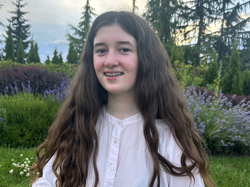A teenage girl with light skin tone, long wavy brown hair and braces stands in a grassy area that has lavender in the background.