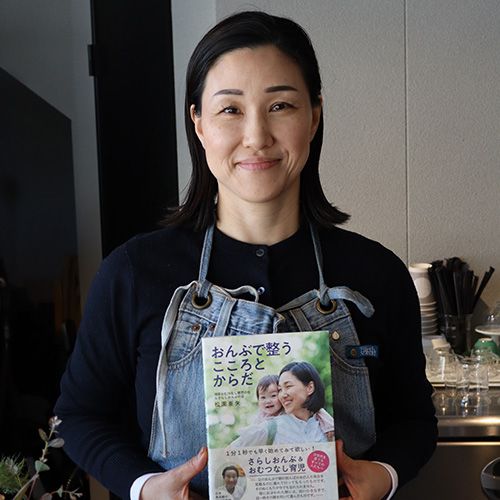 a smiling woman wearing an apron in a cafe holds up a book with a smiling mother and baby on the cover