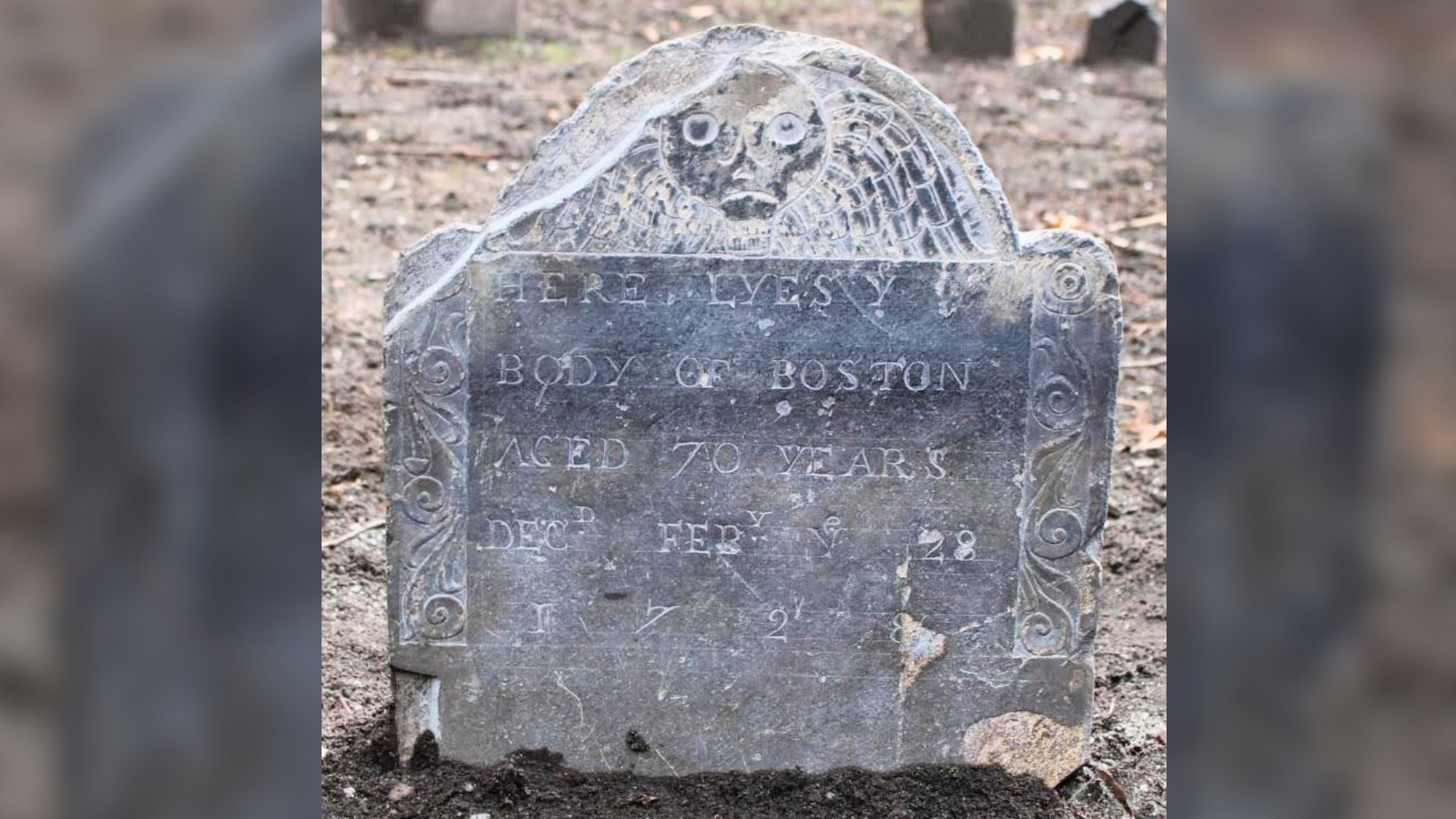 a grey gravestone in a barren landscape with a death's head decoration and inscription