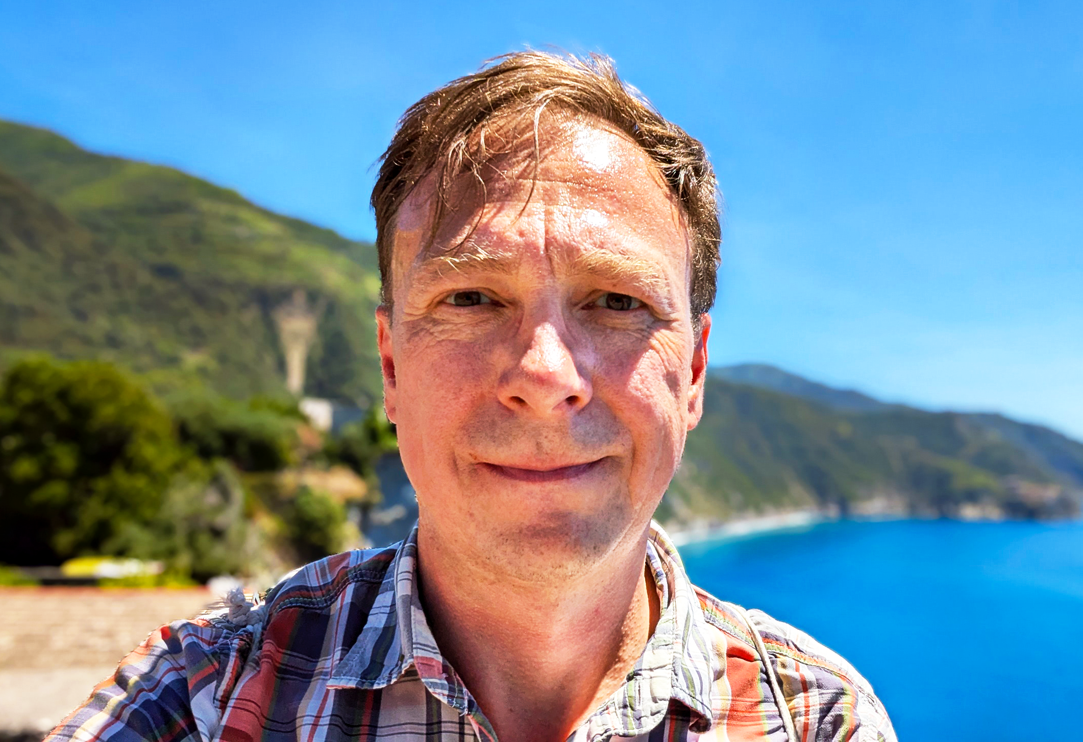 A man wearing a plaid shirt stands outside, in front of a mountain slope meeting the water.