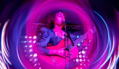 A musician plays guitar and sings into a microphone on stage, surrounded by vibrant pink and purple swirling light effects.