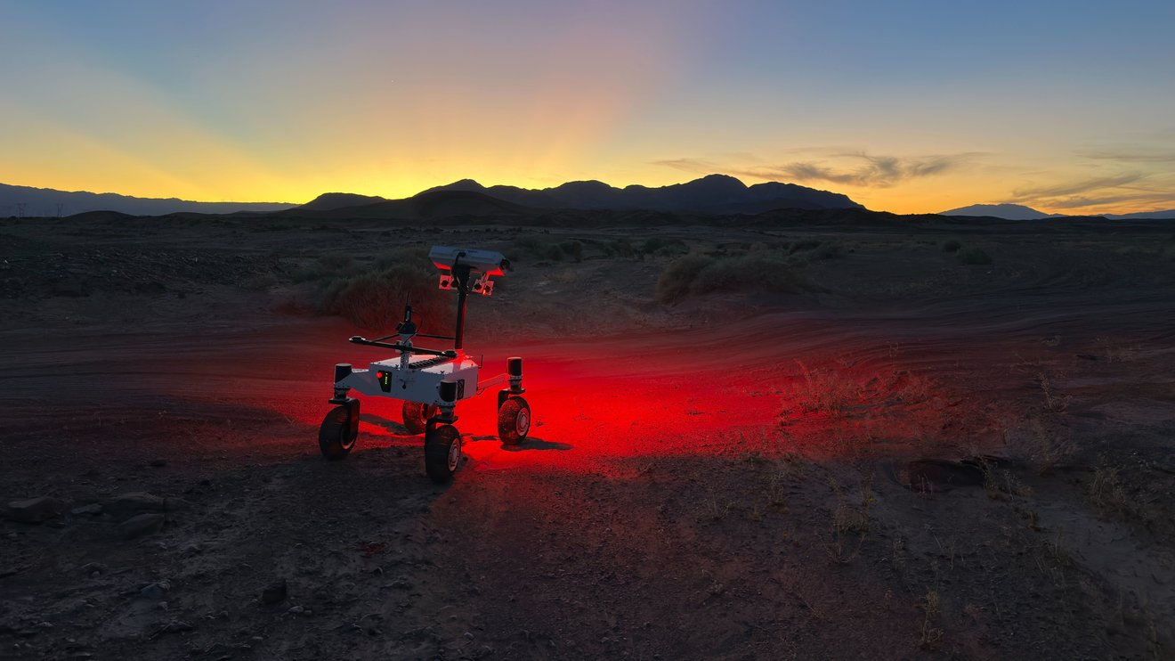 a small, four-wheeled rover shines a red light on a dark desert ground and the sun sets behind silhouetted hills in the background.