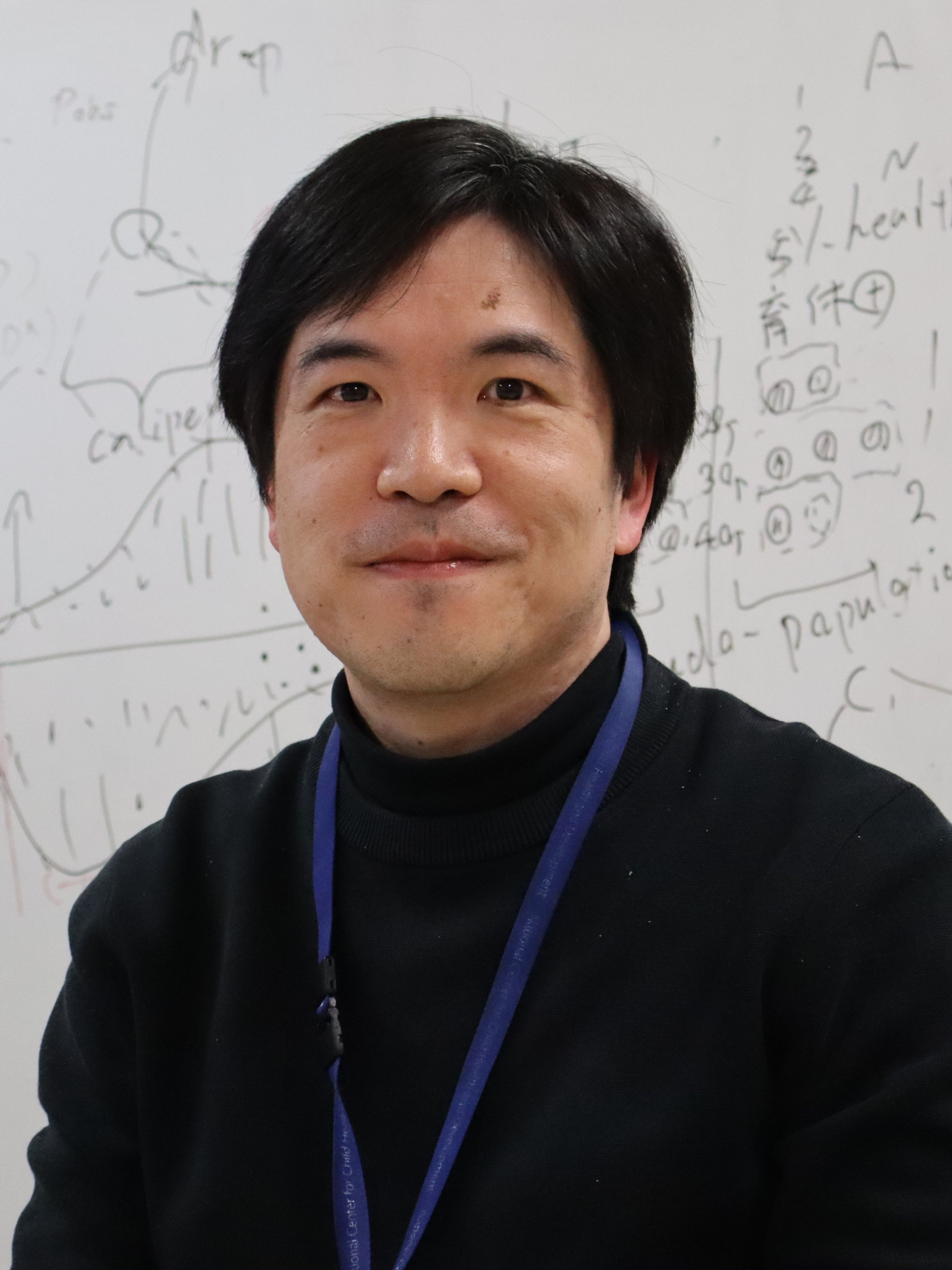 a photo of a smiling man with straight black hair seated in front of a whiteboard with graphs and equations