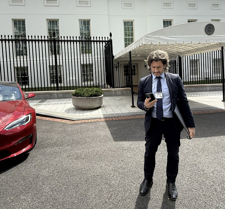 Barron, on the right, looks at his phone. He’s in front of a canopied walkway leading to the West Wing. A red car, to the left, is partially in frame.