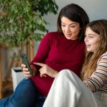 Happy pregnant mother sitting close to smiling teen daughter while taking selfie on smartphone - stock photo